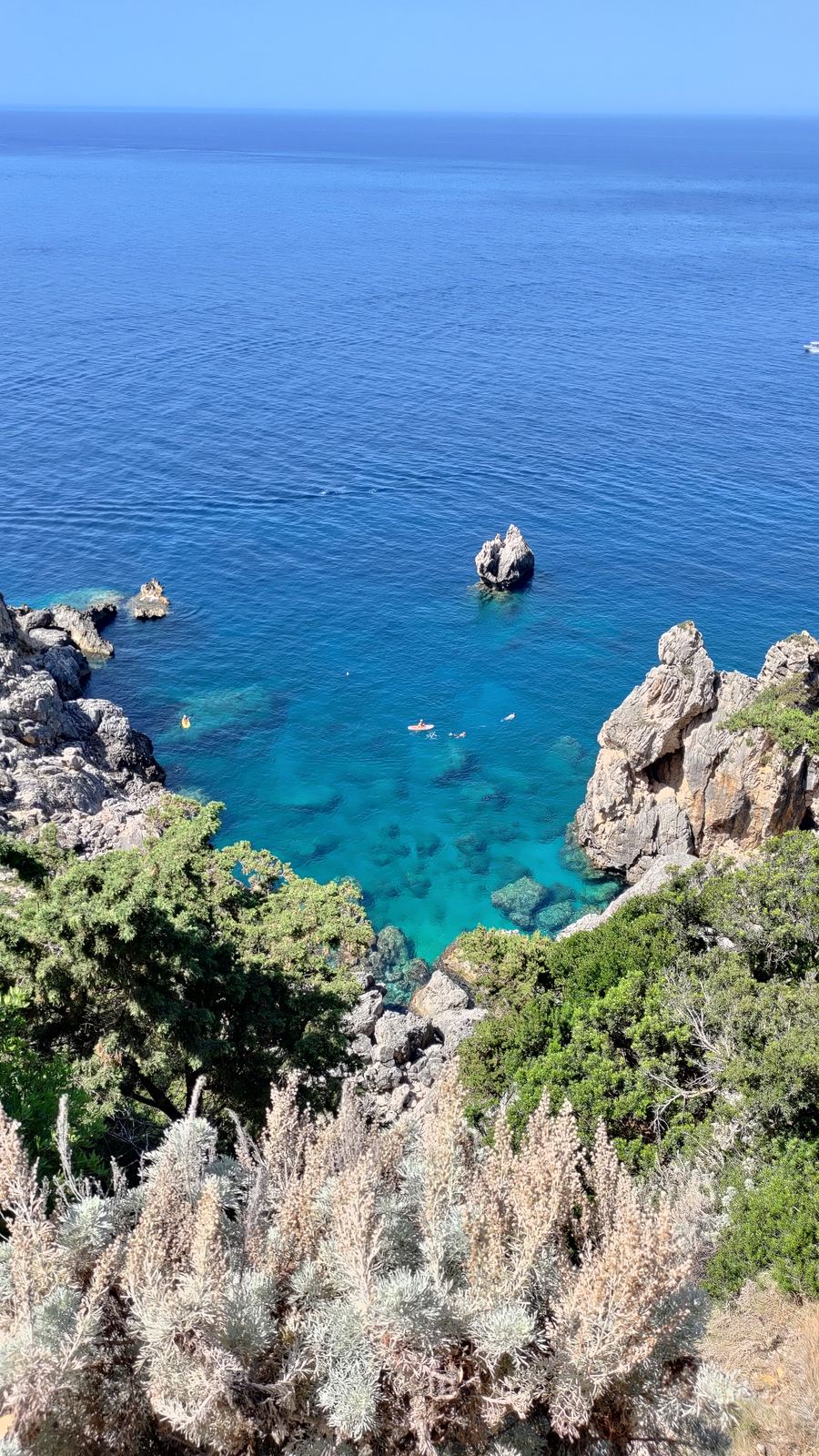 An aerial view of a turquoise cove between rocky, pine-covered cliffs, with small figures on the water.