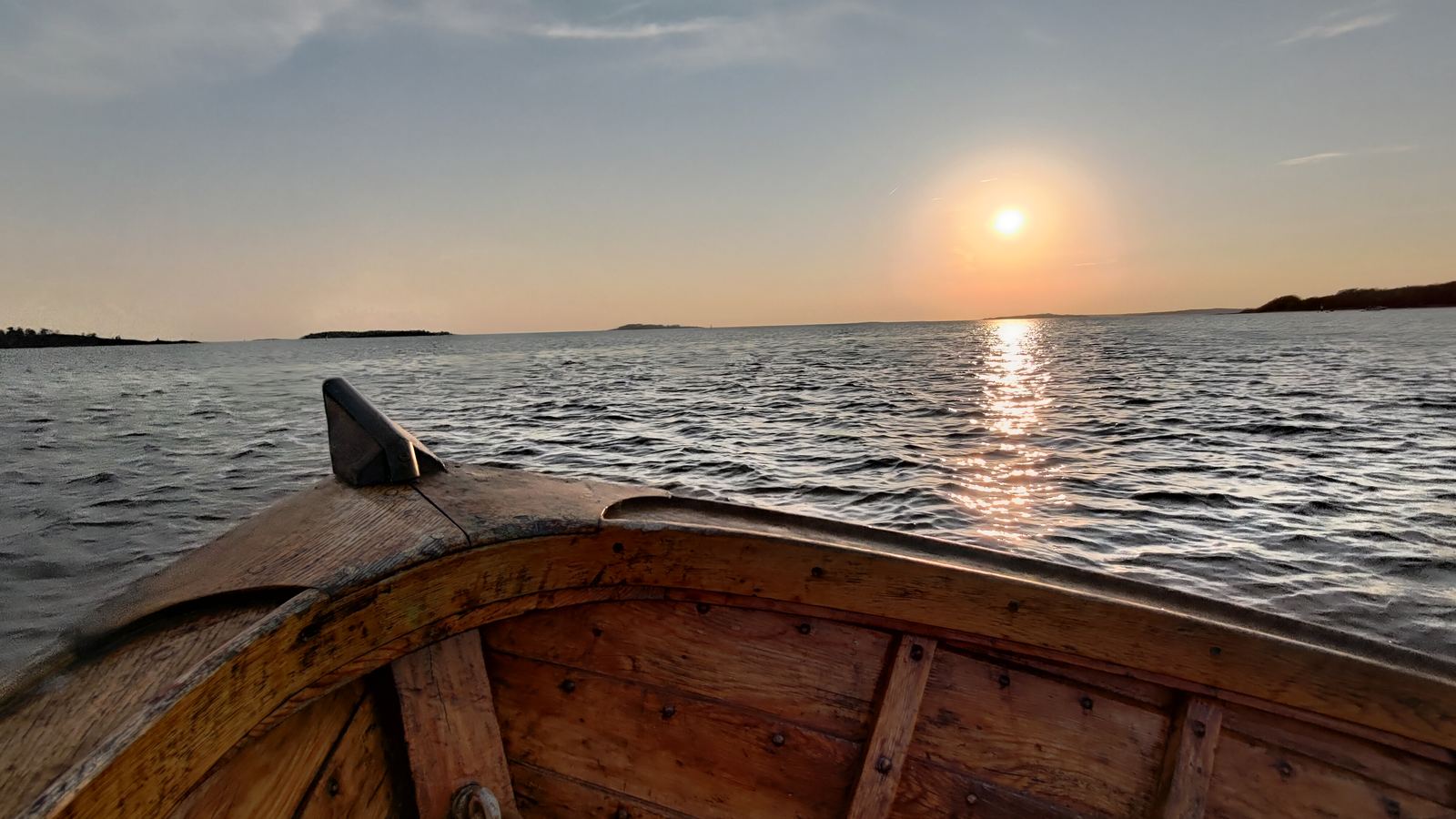 The wooden bow of a rowboat facing a low sun setting over calm water and distant islands.