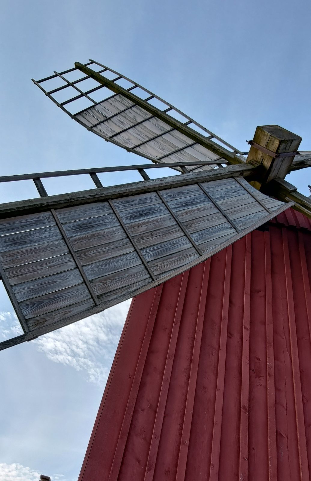 Looking up at the wooden sails of a windmill against a blue sky, next to its red-painted tower.